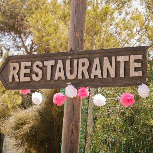 A rustic wooden restaurant sign in Spain, adorned with festive pink and white decorations amidst a natural outdoor setting.